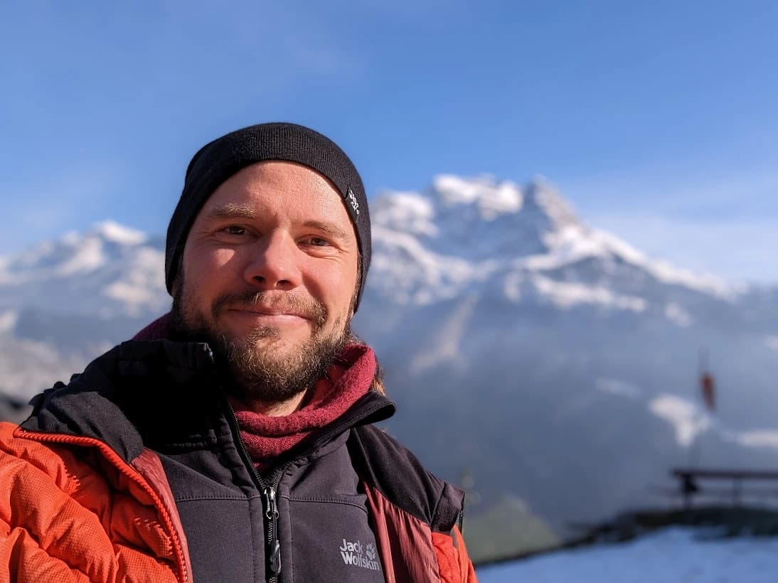 Flight instructor and tandem pilot Miki Mohos smiling, wearing a black beanie and a Jack Wolfskin jacket with snow-covered mountains in the background.